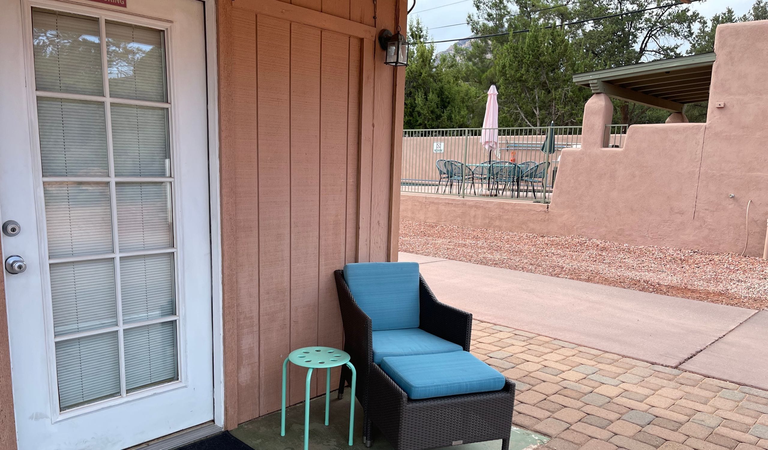 A covered patio area with a wicker chair and a small stool, a door with a window, and outdoor furniture in the background.