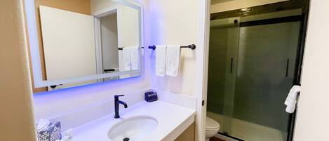 Un baño con lavamanos blanco, espejo, toallero y cabina de ducha con mampara de vidrio.
