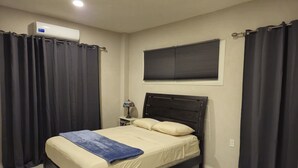 Un dormitorio con cama, mesita de noche, lámpara, ventana con persianas y un equipo de aire acondicionado.