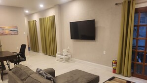 Una sala de estar con un sofá, una mesita, un televisor montado en la pared y una ventana con cortinas.