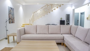 A modern living room with a beige sectional sofa, a wooden coffee table, a staircase with ornate railings, and a painting on the wall. - Badrani A - Qaswarah Residence  (Madinah)