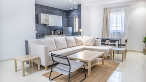 A modern living room with a sectional sofa, a dining table, and a kitchen area with a microwave and cabinets. - Badrani A - Qaswarah Residence  (Madinah)