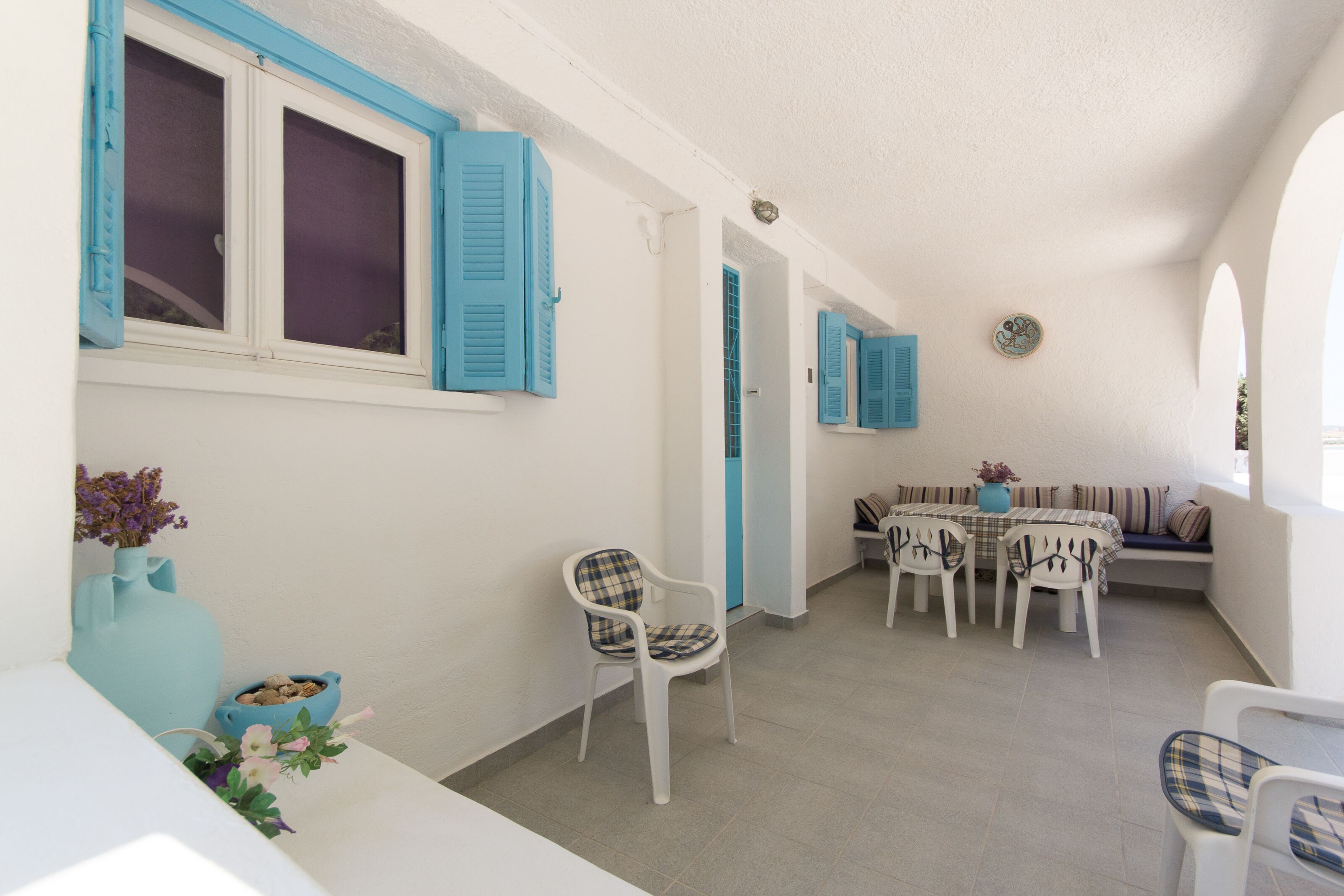 Ein Zimmer mit weißer Wand, einem Fenster mit blauen Fensterläden, einem weißen Tisch mit Stühlen und einer blauen Vase mit Blumen.