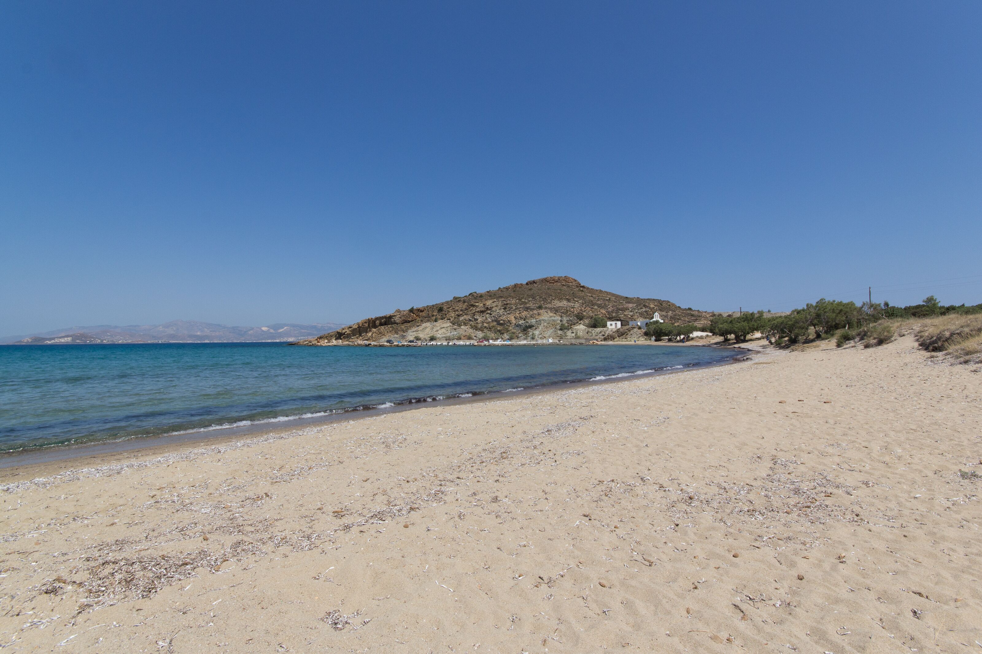Uma praia com areal, uma pequena colina com vegetação rasteira e um céu azul límpido.