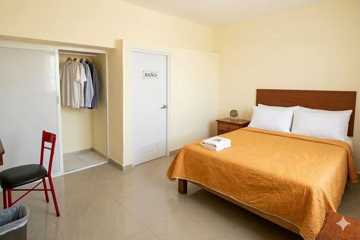 A hotel room with a bed, a chair, a wardrobe, and a door labeled 'BAÑO'.