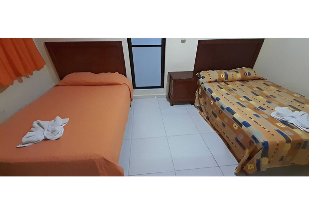 A hotel room with two beds, one with an orange bedspread and the other with a patterned bedspread. There is a small wooden nightstand between the beds.
