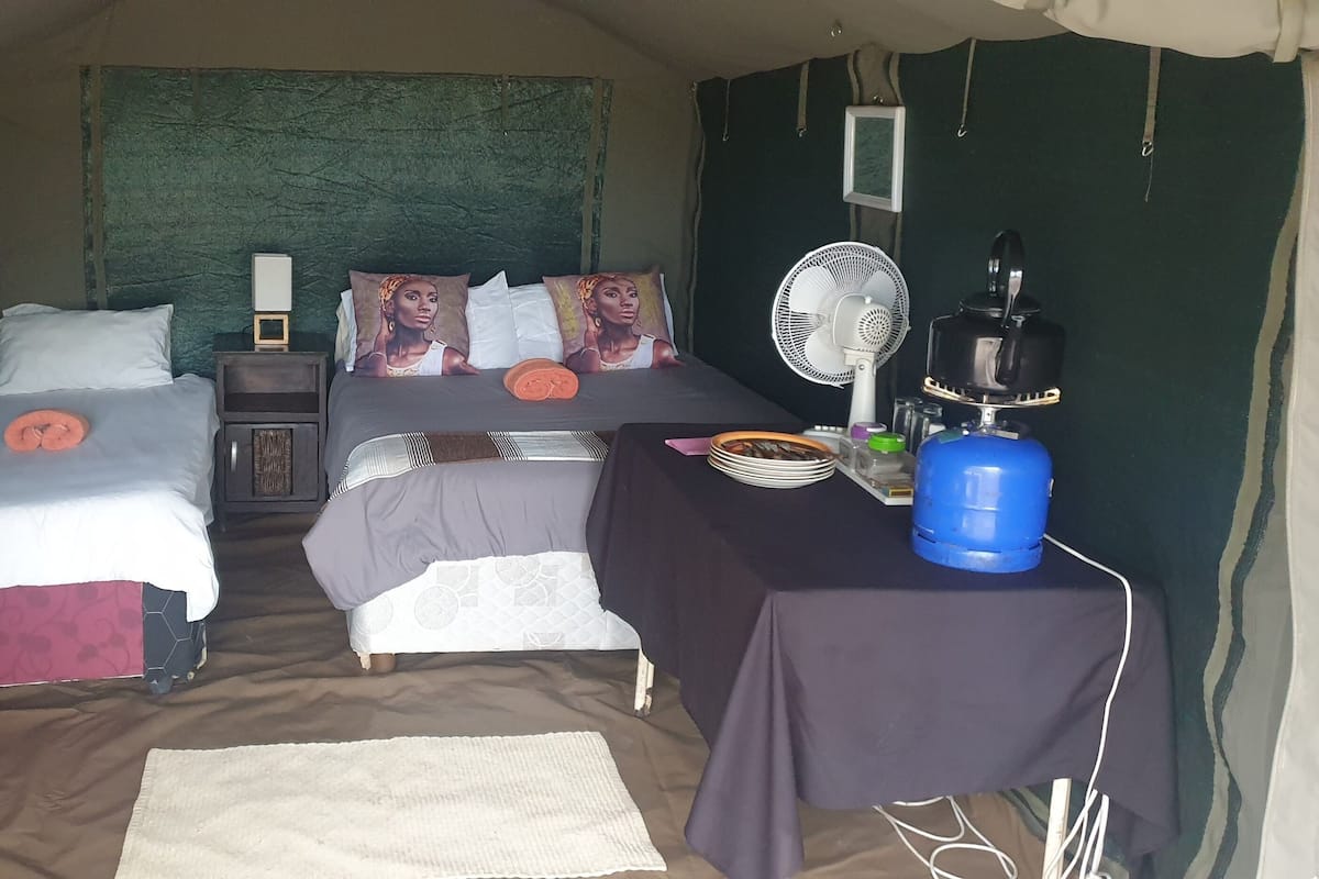 A room with two beds, a table with a kettle and plates, a fan, and a water dispenser.
