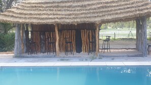 A poolside area with a thatched roof structure, wooden pillars, and metal bar stools.