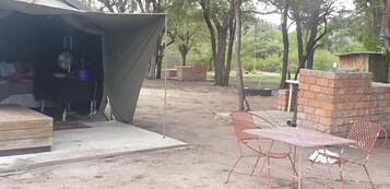A tent with a fan and a water dispenser, a brick structure, and a table with two chairs.