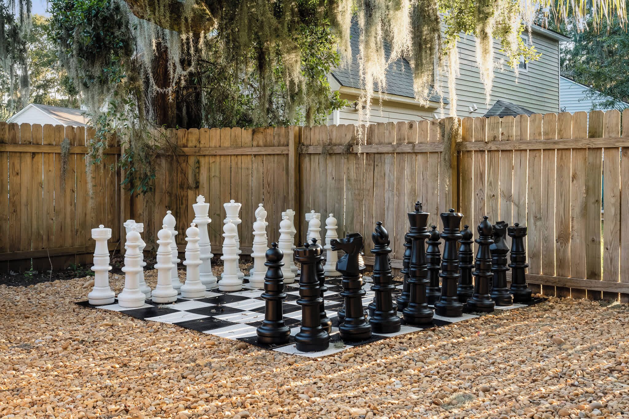 A large outdoor chessboard set up on a gravel surface, surrounded by a wooden fence and trees.