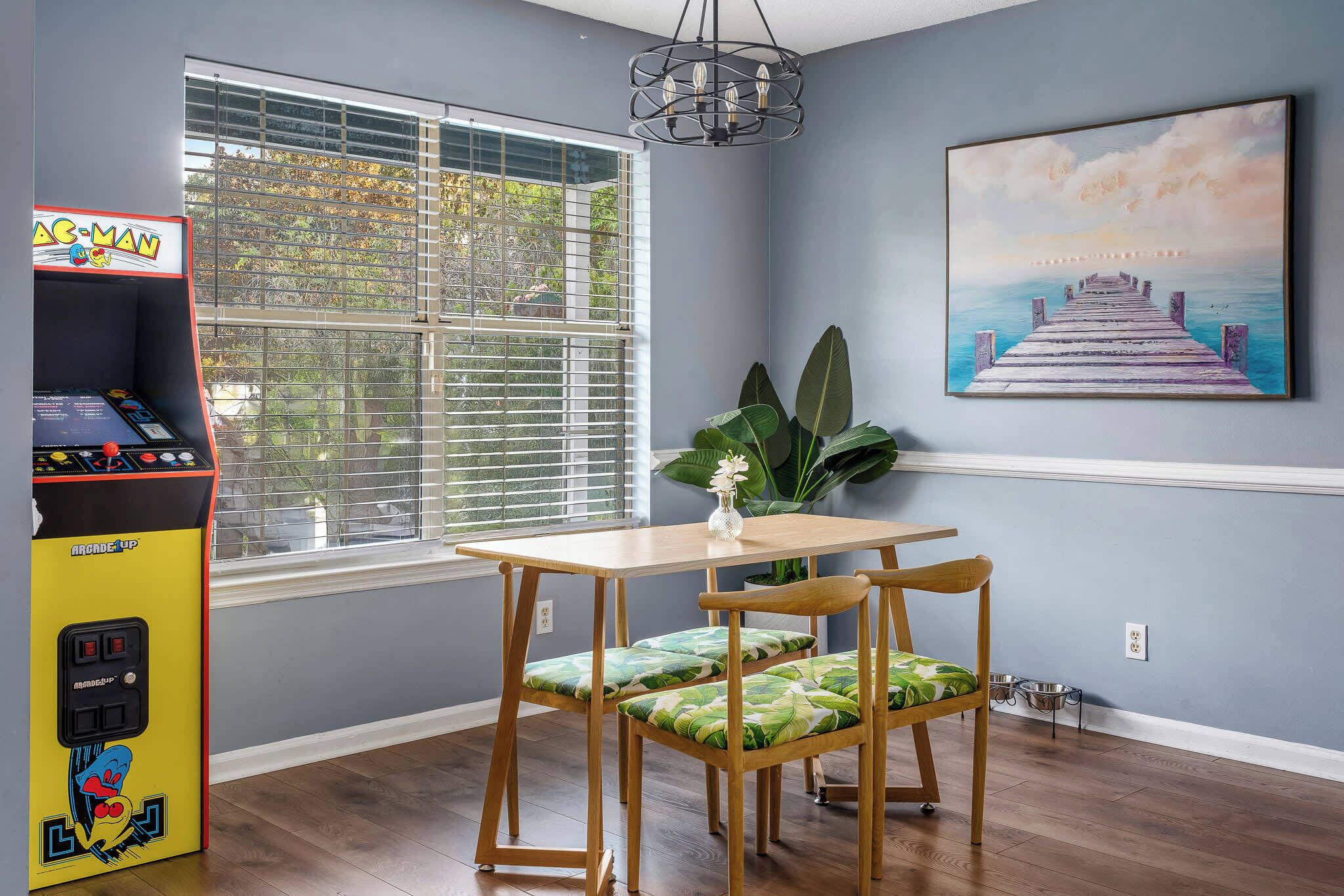 A room with a wooden table and chairs, a window with blinds, a wall-mounted painting, and an arcade machine.
