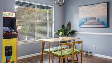 A room with a wooden table and chairs, a window with blinds, a wall-mounted painting, and an arcade machine.