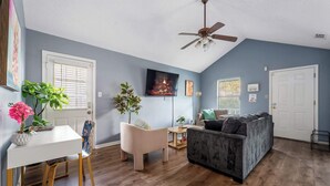Ein Wohnzimmer mit Deckenventilator, einem an der Wand montierten Fernseher, einer Couch, einem Esstisch mit Stühlen und einer Zimmerpflanze.