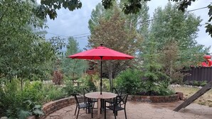 Un espace patio avec une table ronde et des chaises, un parasol rouge et de la verdure tout autour.
