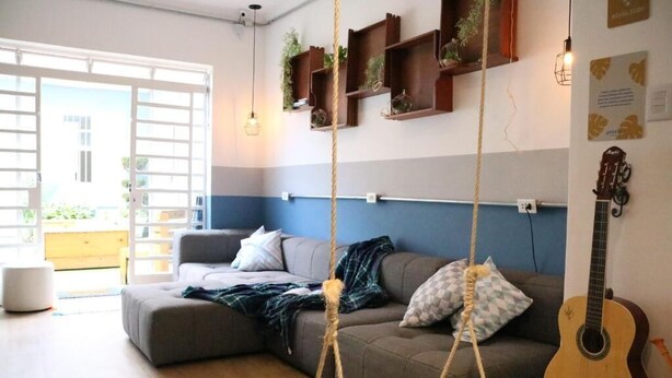 A living room with a grey sectional sofa, a hanging swing, a guitar, and shelves with plants. - Angatu Hostel (São Paulo)