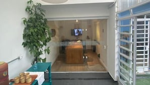 A narrow hallway with a wooden bench and a plant on the left, leading to a room with a TV and a wooden counter. - Angatu Hostel (São Paulo)