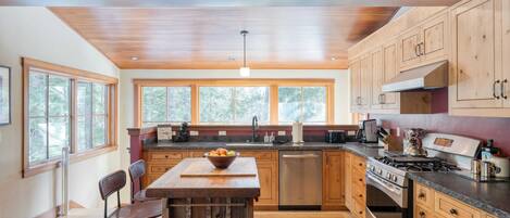 A modern kitchen with wooden cabinets, a central island, stainless steel appliances, and large windows providing natural light.