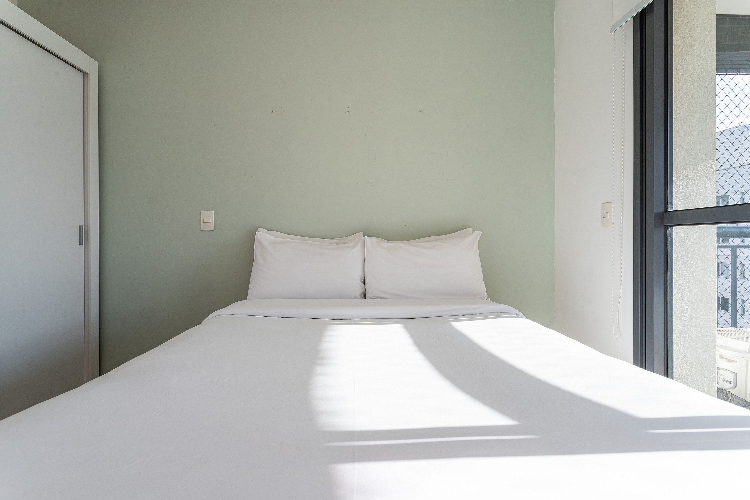Un lit avec une literie blanche dans une pièce dont un mur est peint en vert clair et qui possède une fenêtre.