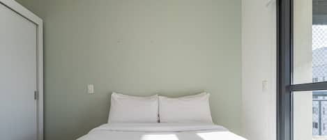 A bed with white bedding in a room with a light green wall and a window.