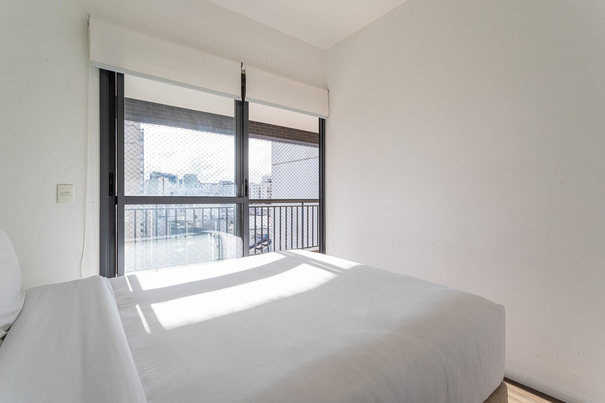 A hotel room with a large bed, a window with a view of the city, and a balcony with a railing.