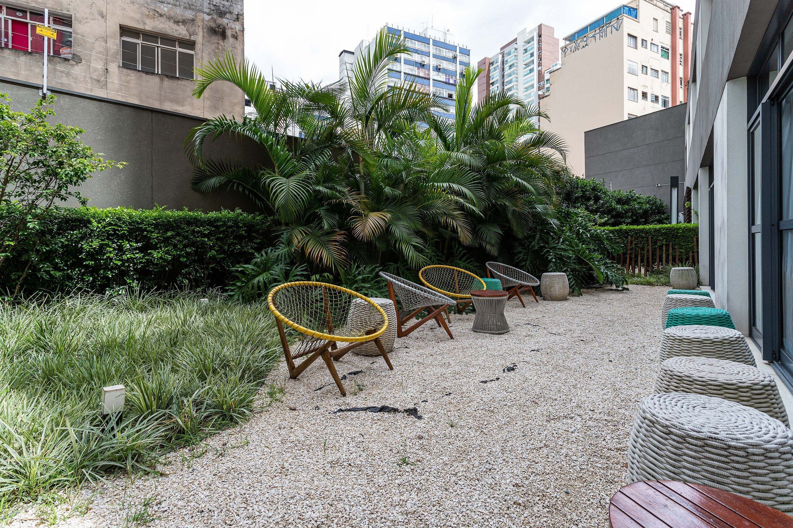 A courtyard with outdoor seating, including wicker chairs and a wooden table, surrounded by lush greenery and a gravel path.