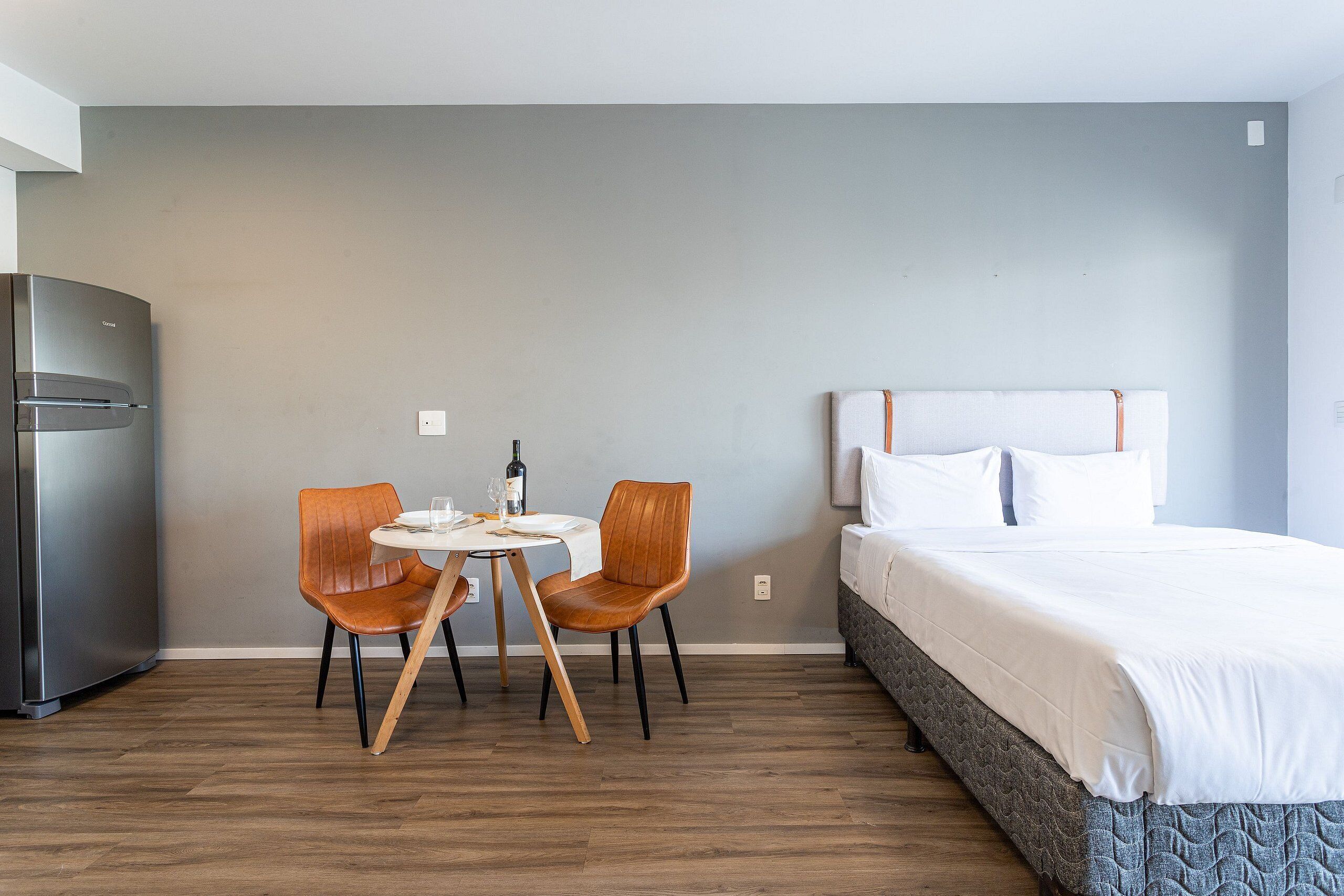 A hotel room with a bed, a dining table with two chairs, a refrigerator, and a wall with a light switch.