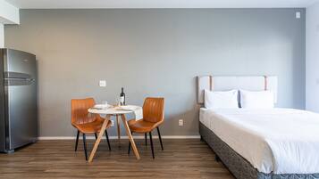 A hotel room with a bed, a dining table with two chairs, a refrigerator, and a wall with a light switch.