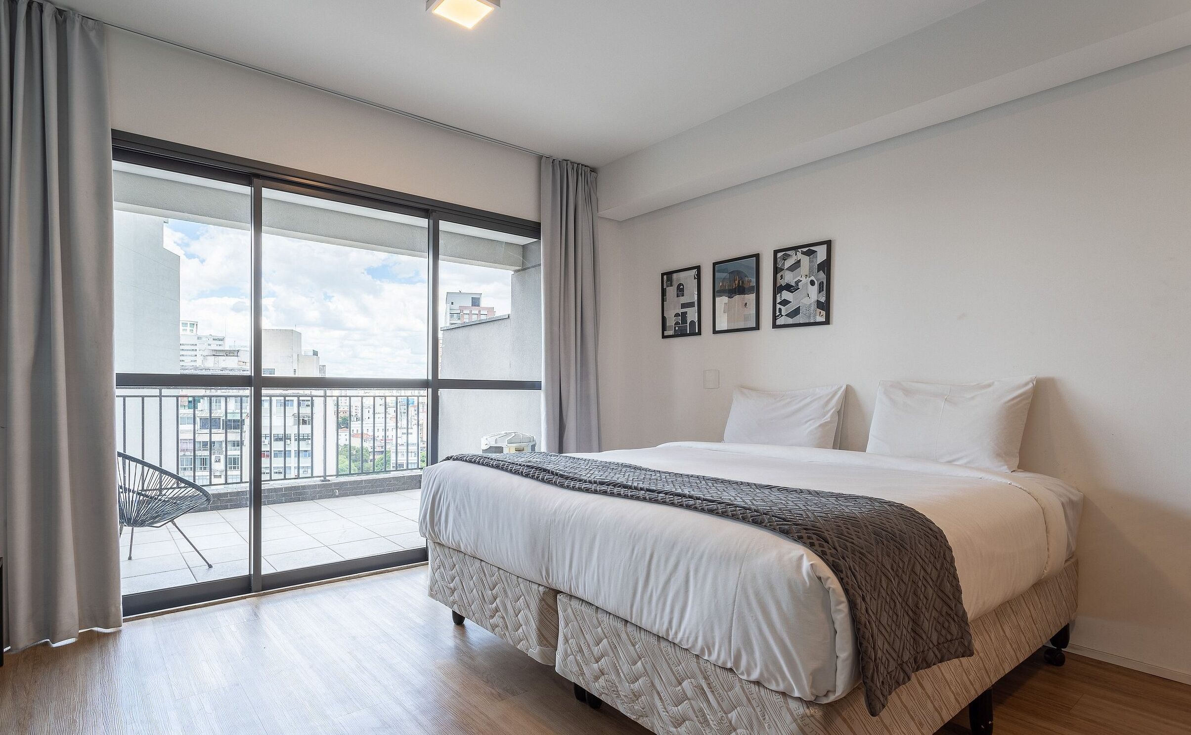A bedroom with a large bed, a view of the city through a sliding glass door, and three framed pictures on the wall.