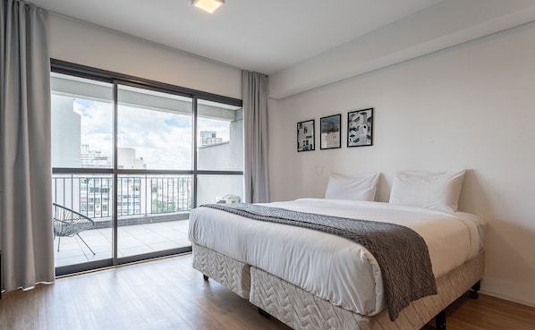 Quarto com uma cama grande, vista da cidade através de uma porta de correr de vidro e três quadros na parede.