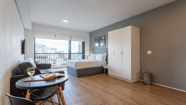 A modern hotel room with a bed, a seating area with a table and chairs, a wardrobe, and a view of the city through large windows. - Setin Downtown Genebra by Viva - Apartamentos completos na Bela Vista com piscina (São Paulo)