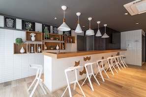 Uma área de bar moderna com banquetas brancas, prateleiras de madeira com plantas e iluminação pendente.