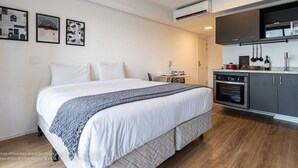 A modern bedroom with a large bed, a small table, and a kitchenette area. - Setin Downtown Genebra by Viva - Apartamentos completos na Bela Vista com piscina (São Paulo)