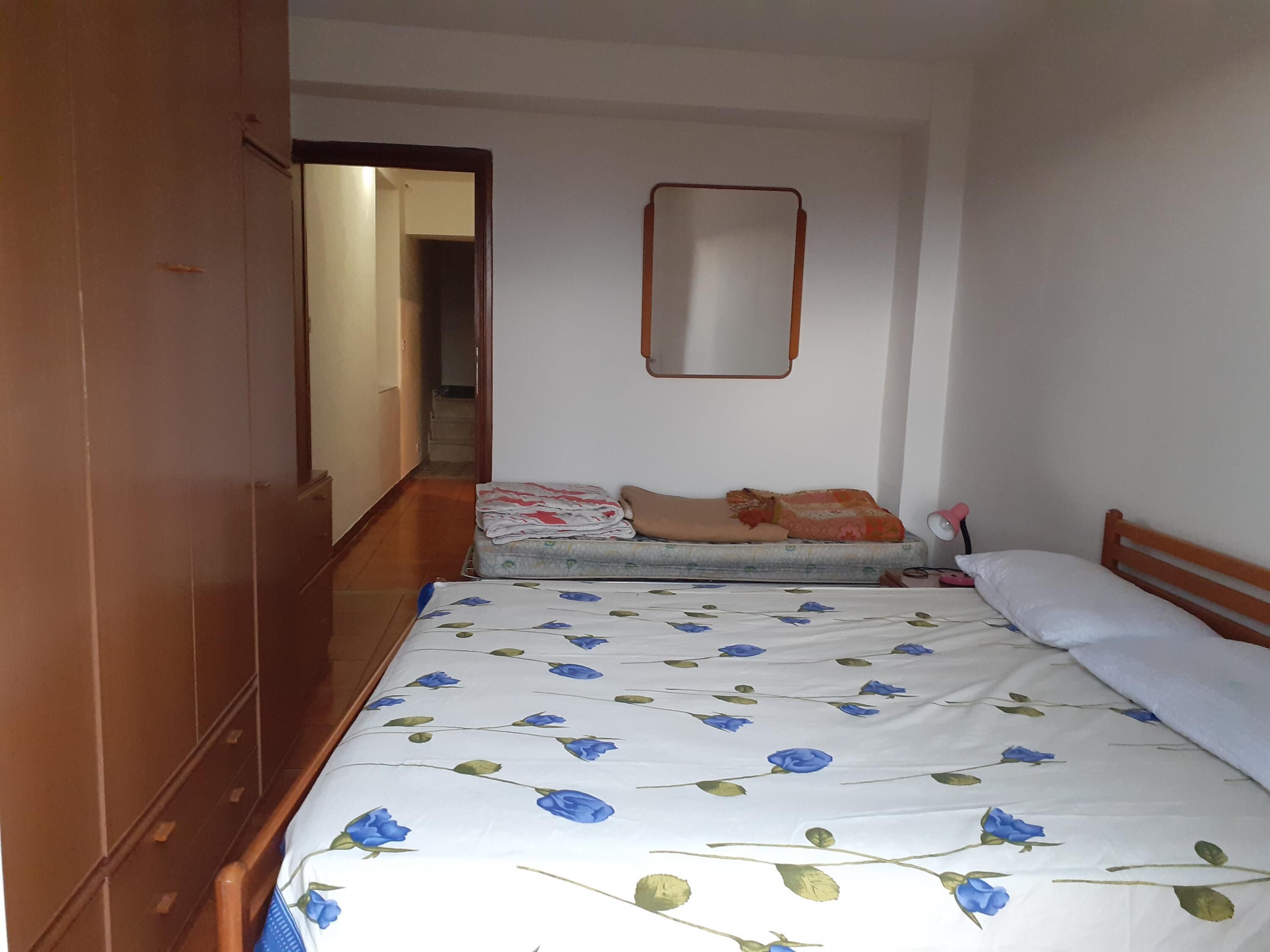 Un dormitorio con un armario de madera, una cama con colcha estampada, una mesita de noche con lámpara y un espejo en la pared.