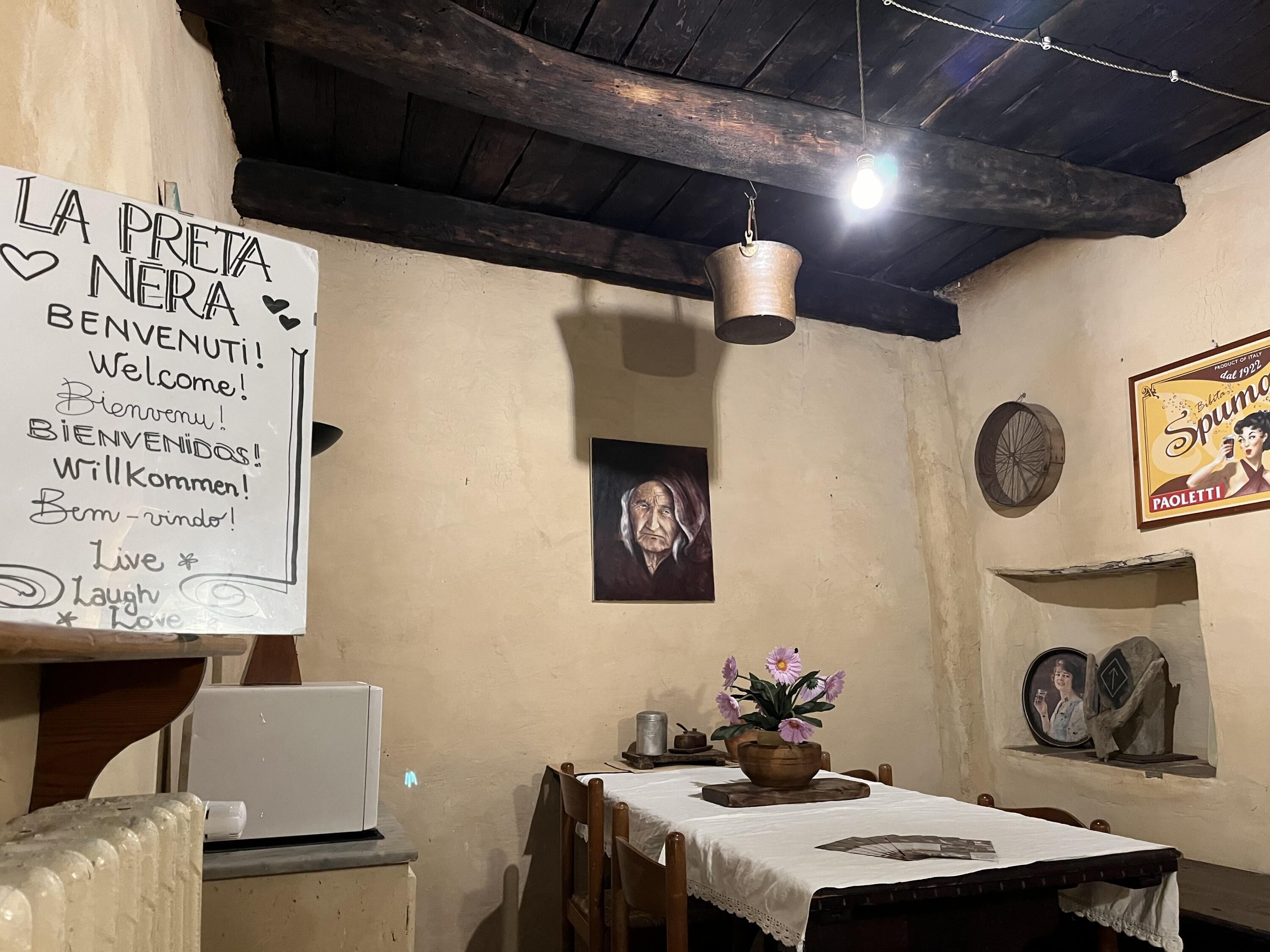 A rustic dining area with a wooden table, a white tablecloth, and a vase of flowers. A sign on the wall reads "LA PRETA NERA" with various welcome phrases.