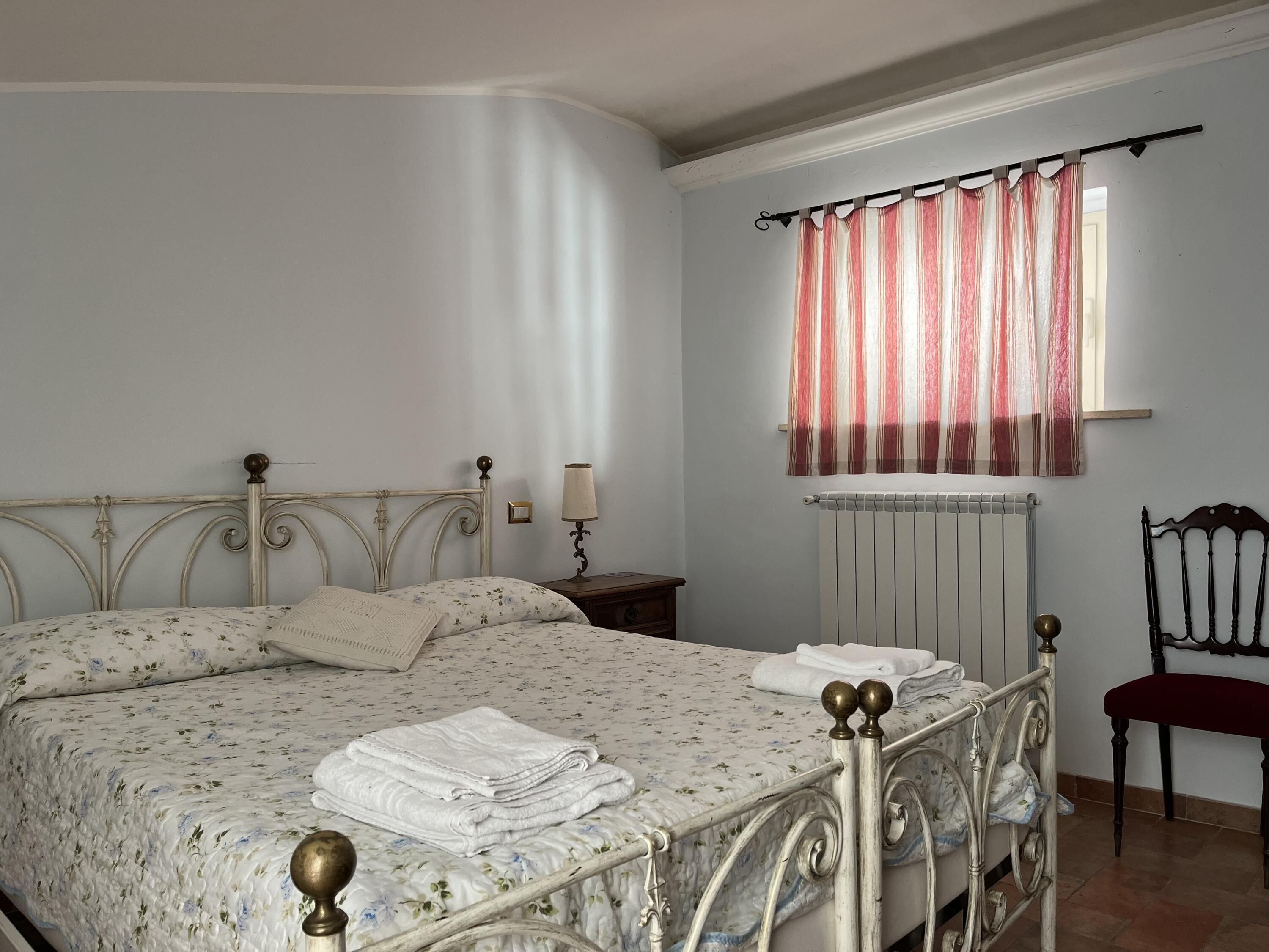 Un dormitorio con cama, mesita de noche, lámpara, silla y ventana con cortinas.