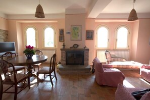Ein Wohnzimmer mit Kamin, Esstisch und Stühlen. Zwei Fenster mit Rundbögen und ein Fernseher links.