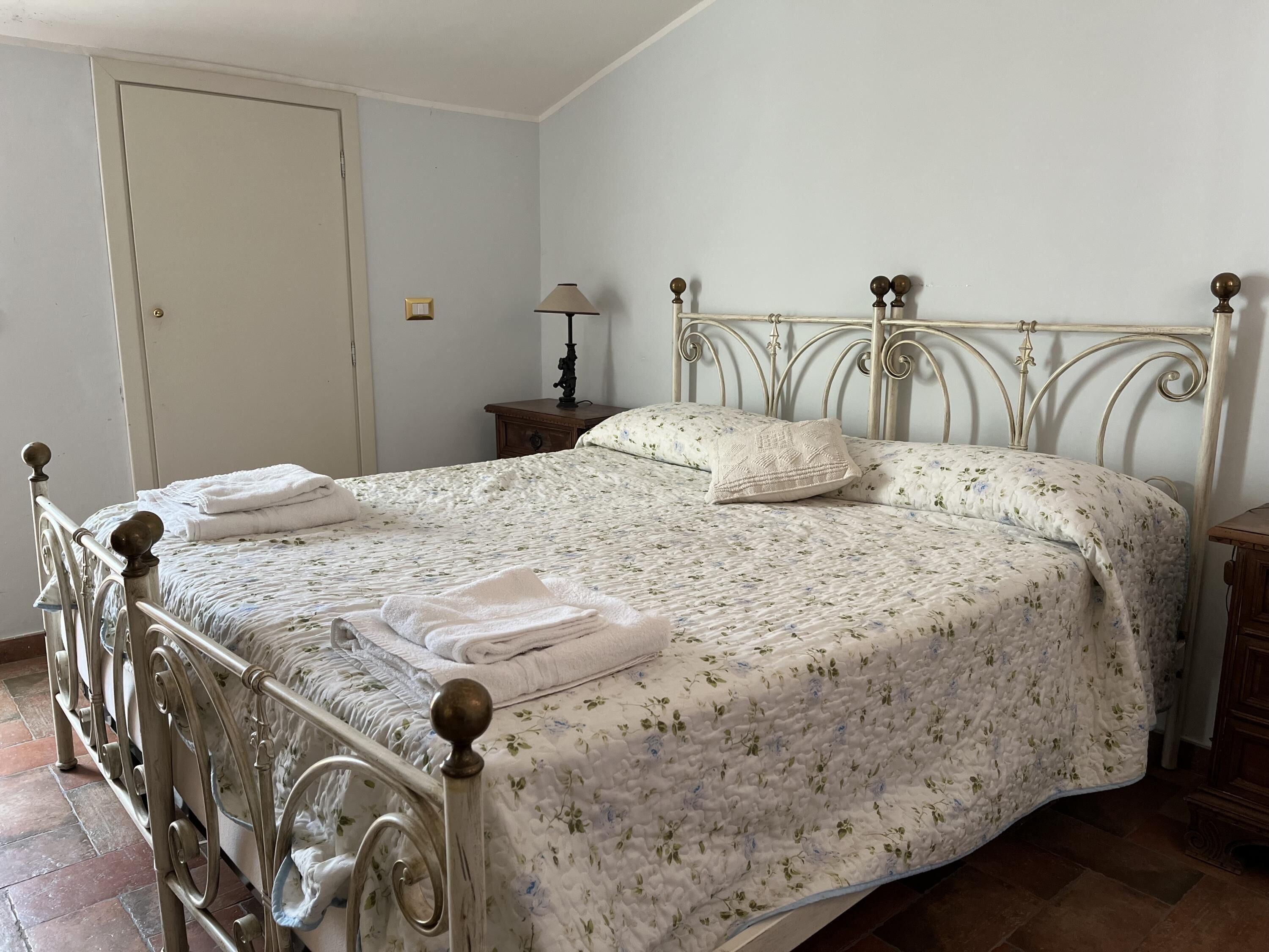 Een net opgemaakt bed met een bloemenprint sprei en twee kussens, een nachtkastje met een lamp, en een deur op de achtergrond.