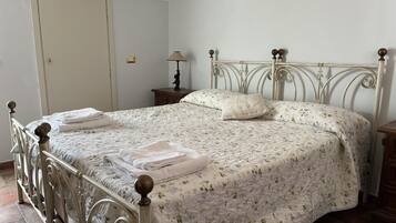Een net opgemaakt bed met een bloemenprint sprei en twee kussens, een nachtkastje met een lamp, en een deur op de achtergrond.