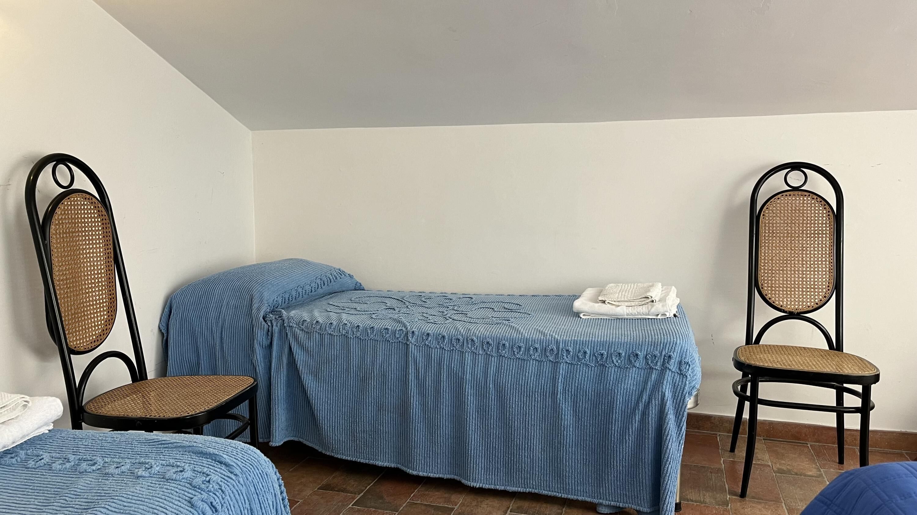 A small, sparsely furnished room with two single beds, two wicker chairs, and a small table.
