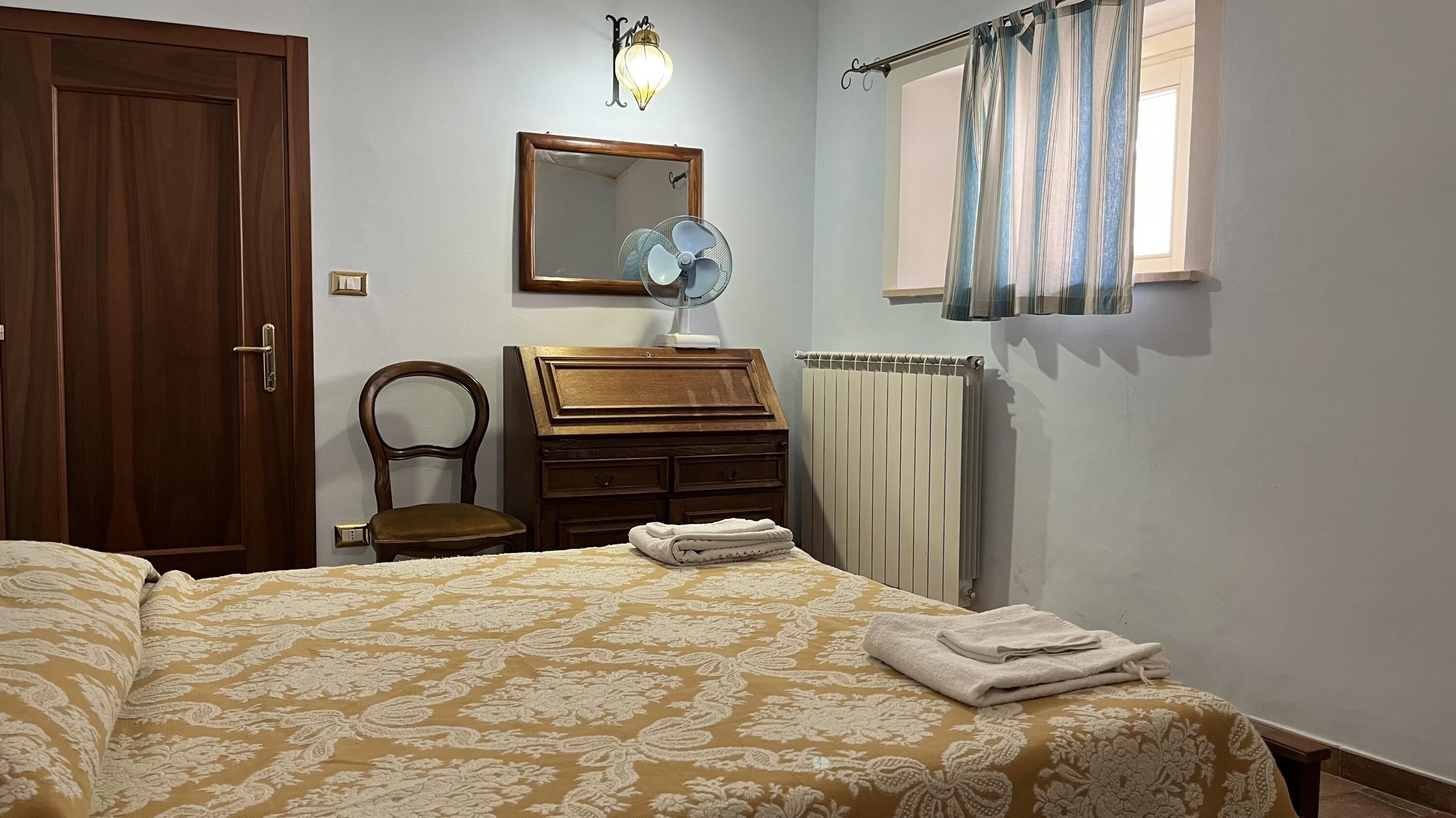 A bedroom with a bed, a wooden dresser, a chair, a mirror, a radiator, and a window with curtains.