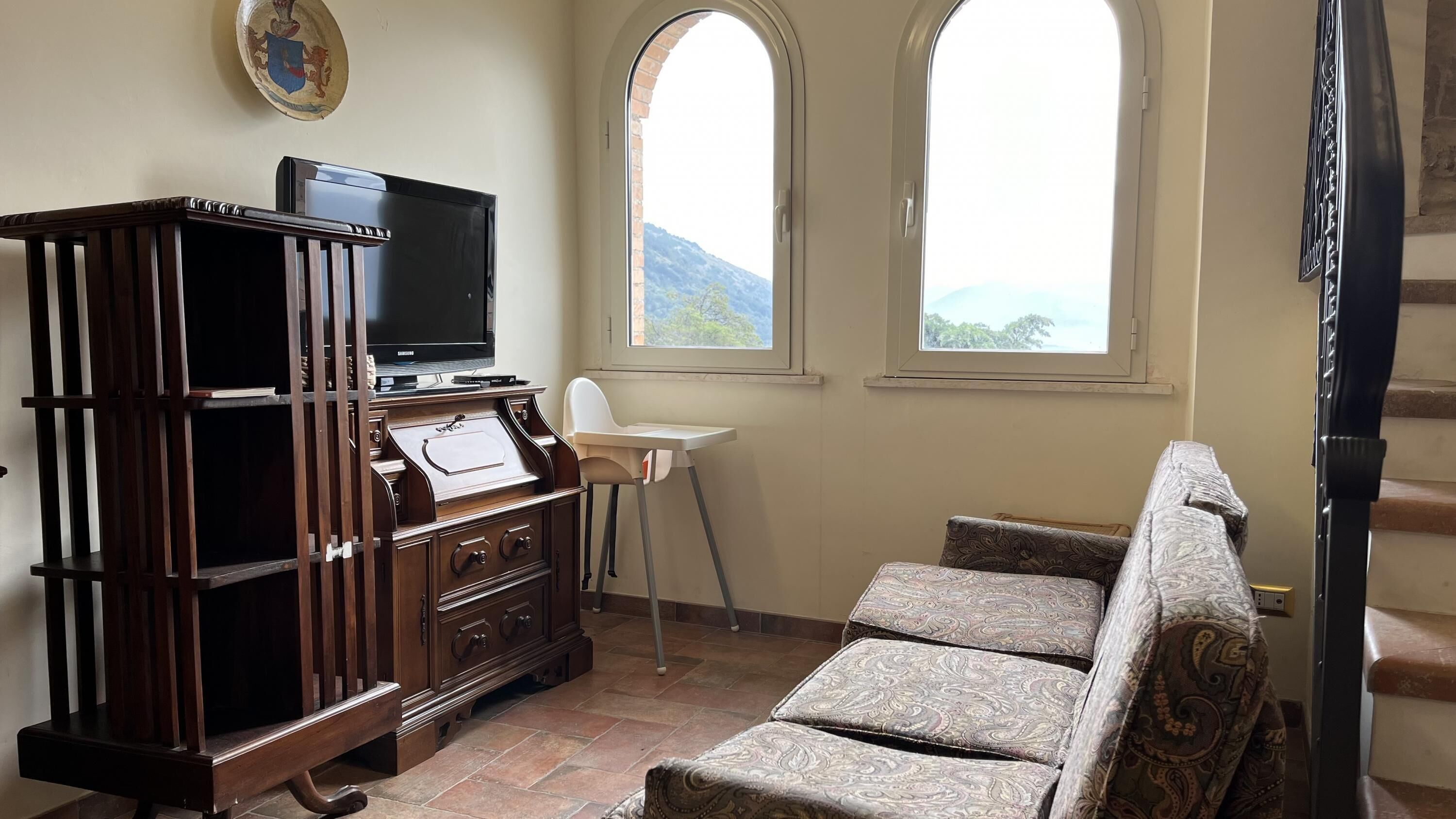 A living room with a wooden entertainment center, a television, a high chair, and a sofa. There are two windows with a view of a mountain.