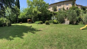 A grassy backyard with a play area, including a slide and climbing structure, surrounded by trees and a stone building in the background. - Villa Felici Montecristo (Giuliano di Roma)