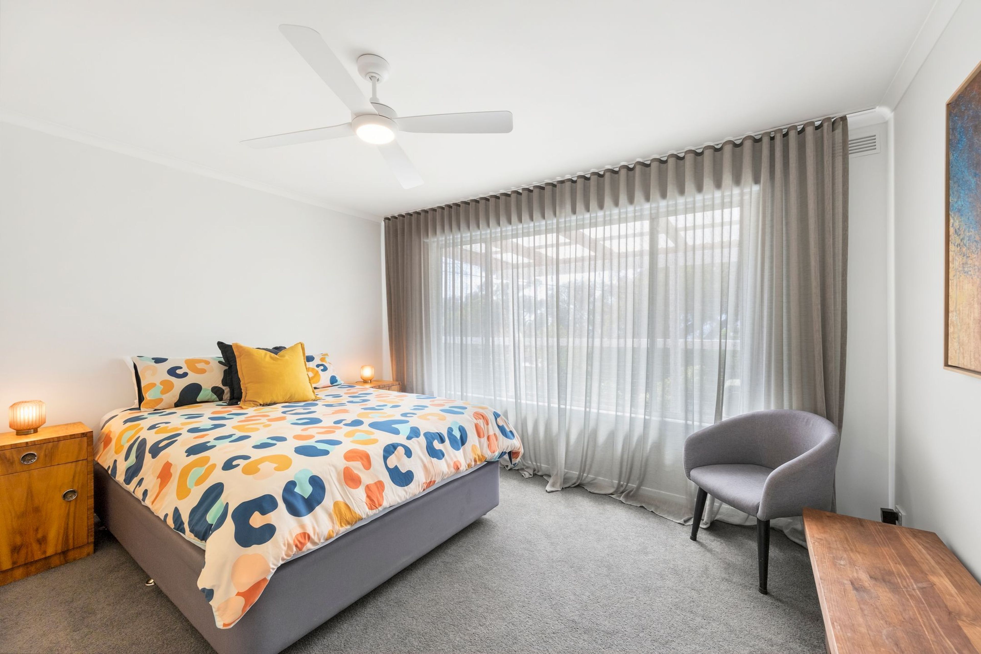 A bedroom with a bed, a chair, a nightstand, a ceiling fan, and a window with curtains.