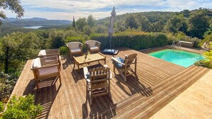 Una terraza de madera con mobiliario exterior, una piscina y una vista panorámica de colinas y árboles.