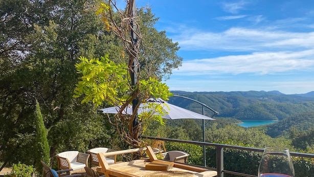 Una terraza de madera con mobiliario exterior, una mesa con un plato y una copa de vino tinto, y vistas a un lago rodeado de colinas.