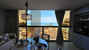 A room with a view of the beach, a dining table set with plates and glasses, a hanging light fixture, and a sofa. - Flat FM20 Cabo Branco Light - By Almare (João Pessoa)