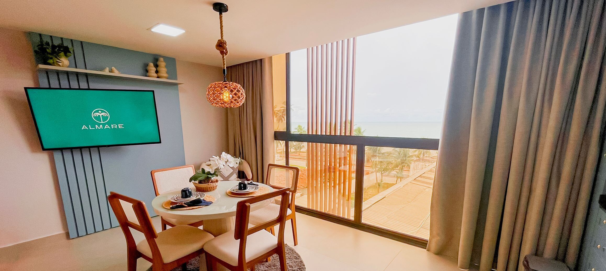 Un espace repas moderne avec une table ronde, des chaises, un téléviseur fixé au mur et une vue sur l’océan grâce à de grandes fenêtres.