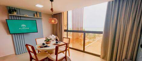 Un espace repas moderne avec une table ronde, des chaises, un téléviseur fixé au mur et une vue sur l’océan grâce à de grandes fenêtres.