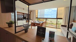 A modern hotel room with a dining area, a kitchen, and a large window offering a view of palm trees and a road. - Flat FM20 Cabo Branco Light - By Almare (João Pessoa)