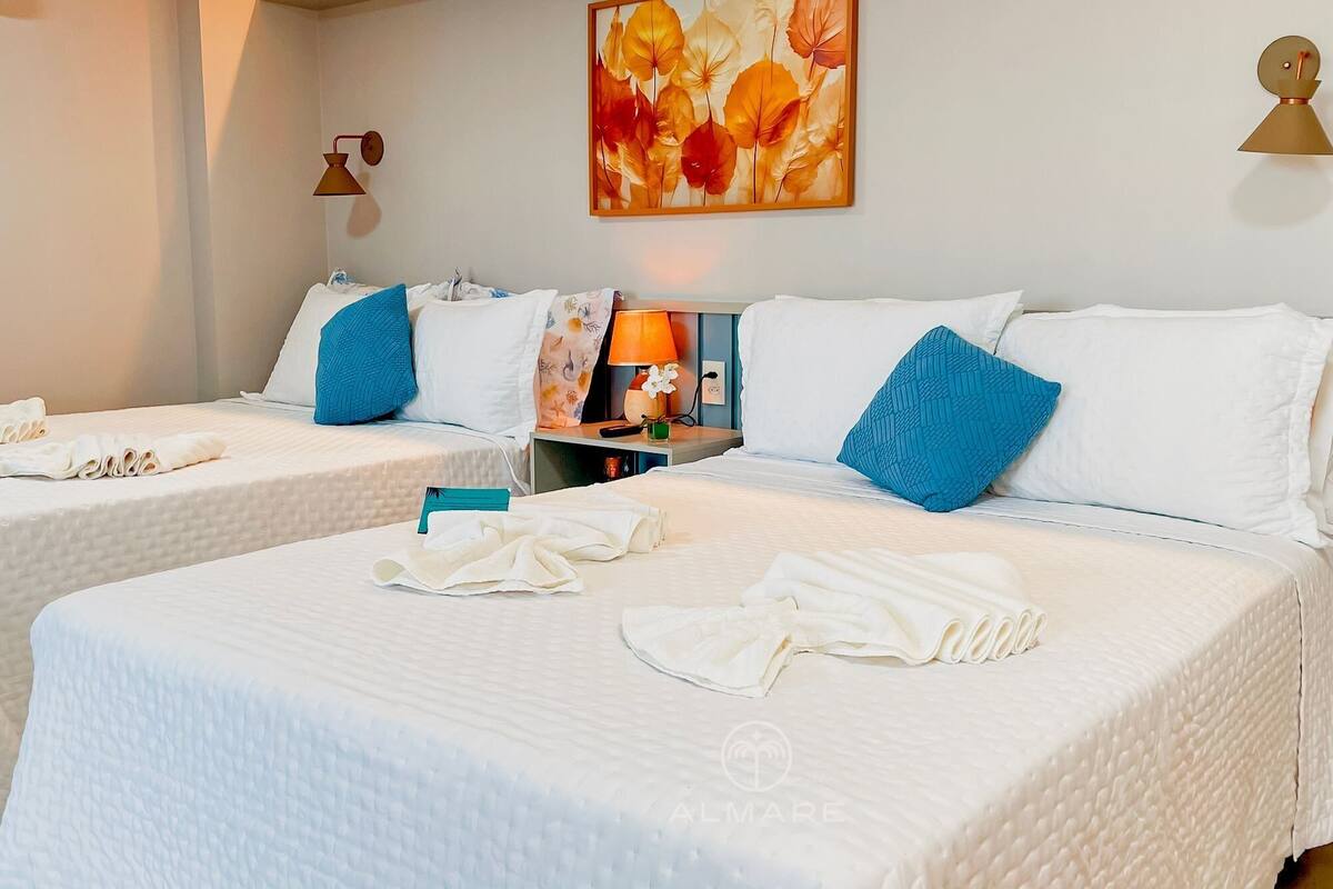 Une chambre d’hôtel avec deux lits, chacun agrémenté de draps blancs et d’oreillers bleus. Un tableau est accroché au mur, une lampe se trouve sur la table de chevet, et une petite table porte un vase avec une fleur.